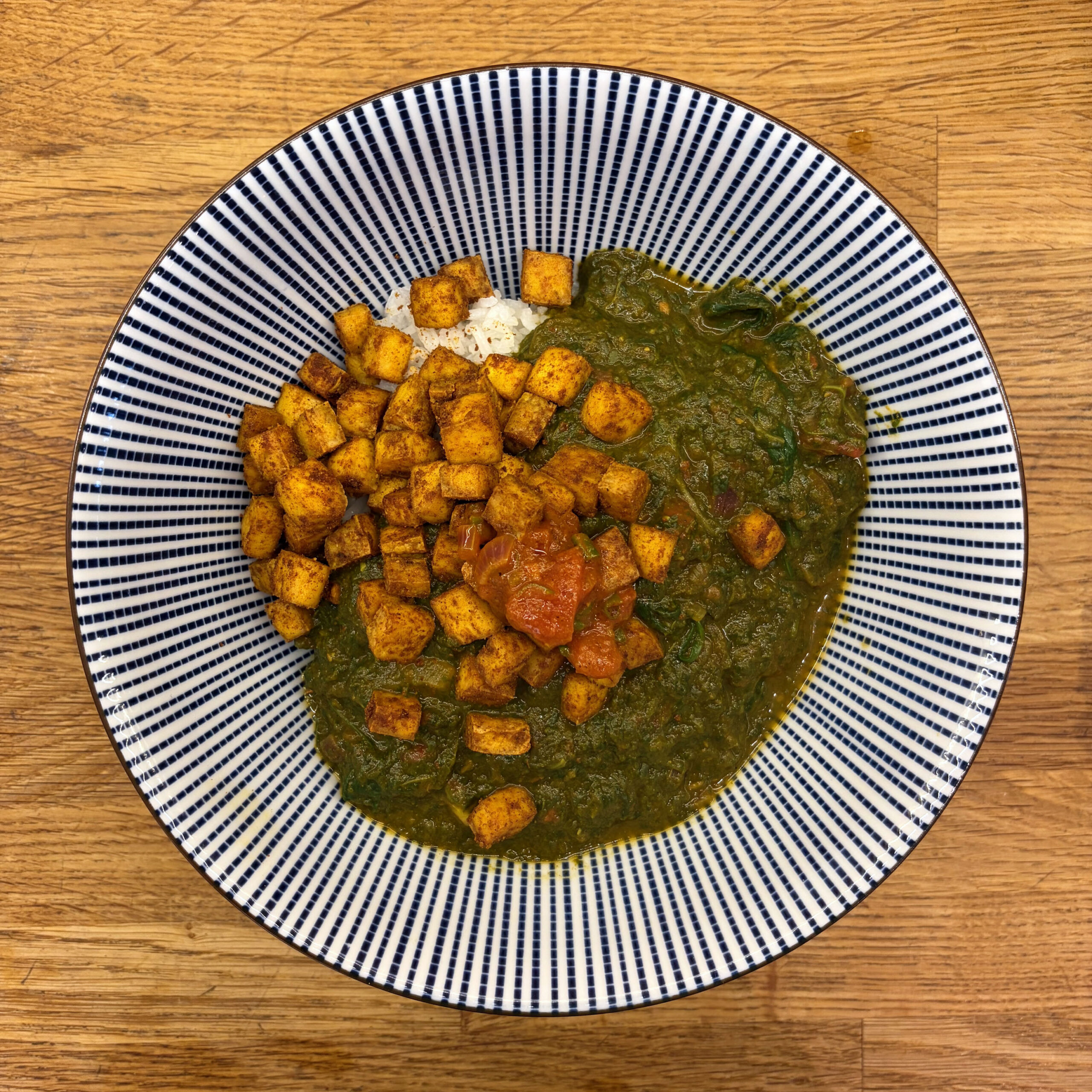 Ein Teller mit Palak Tofu: gewürfelter, mit Gewürzen gerösteter Tofu auf cremigem Spinatcurry mit Tomatenstücken, dazu eine kleine Portion weißer Basmatireis. Serviert in einer blau-weiß gestreiften Schüssel auf einem Holztisch.