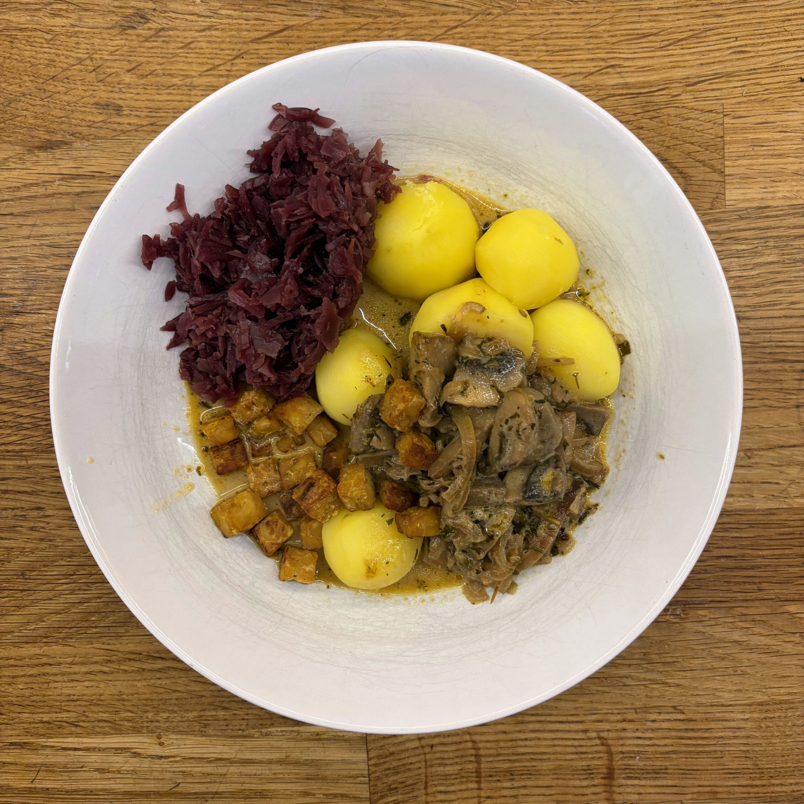 Teller mit Pilzragout aus Champignons in heller Sahnesauce, kleinen ganzen Salzkartoffeln, glasierten Kichererbsen-Tempeh-Würfeln und geschmortem Rotkohl.