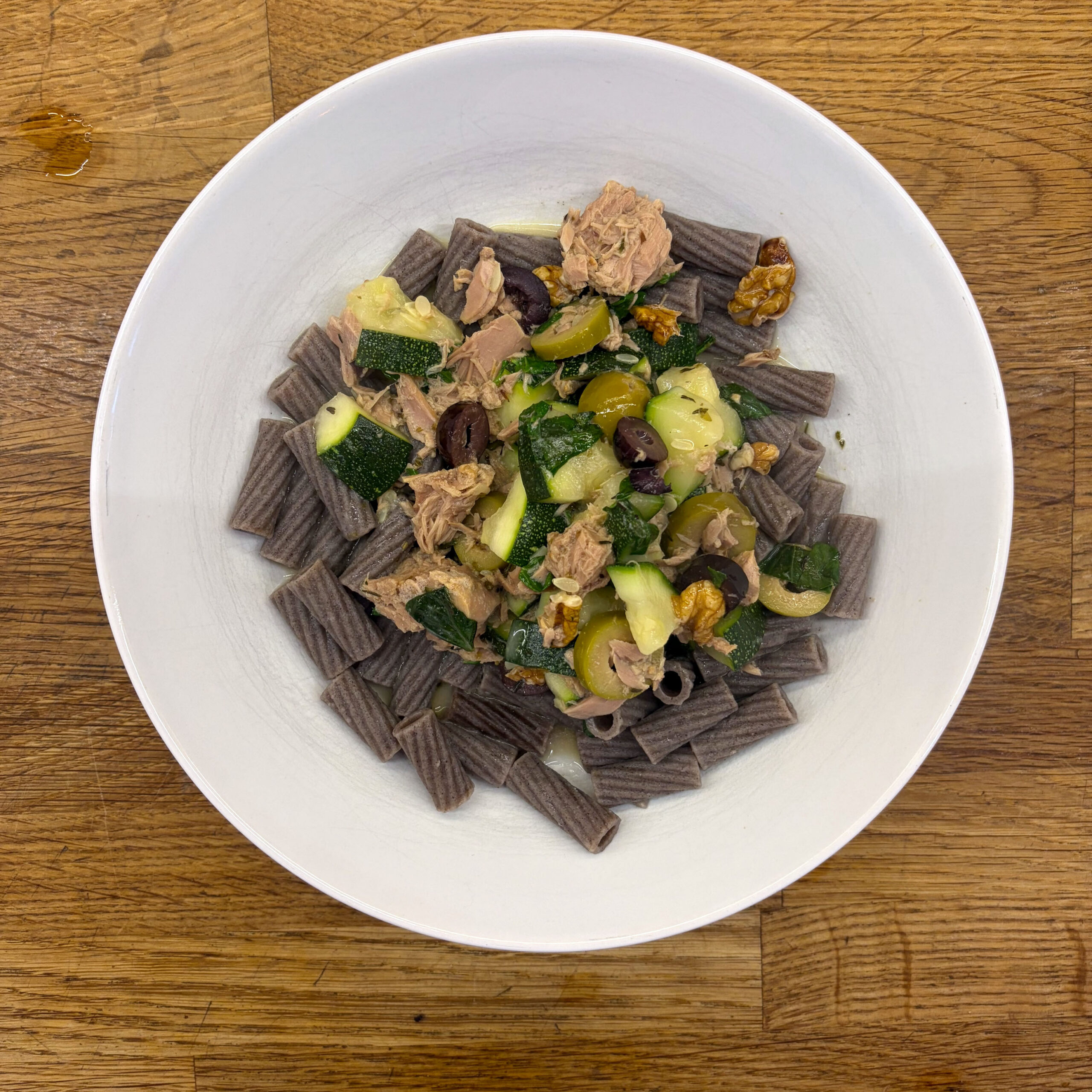 Weißer tiefer Teller mit grauen Buchweizennudeln (Rigatoni-Form), belegt mit einer Thunfisch-Zucchini-Soße mit grünen und schwarzen Oliven sowie Walnüssen, serviert auf einem Holztisch.