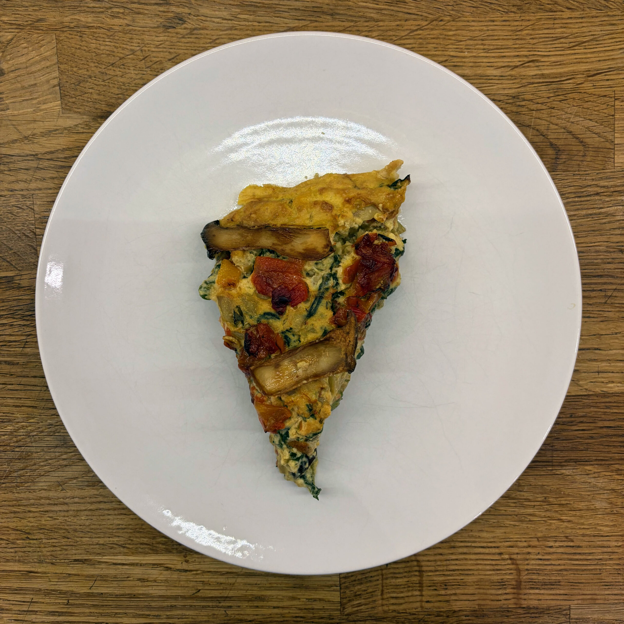 Ein Stück vegane Mangold-Tarte mit sichtbaren Kräuterseitlingen, gegrillter roter Paprika und gehacktem Mangold in einer goldbraunen Füllung auf einem weißen Teller, von oben fotografiert.