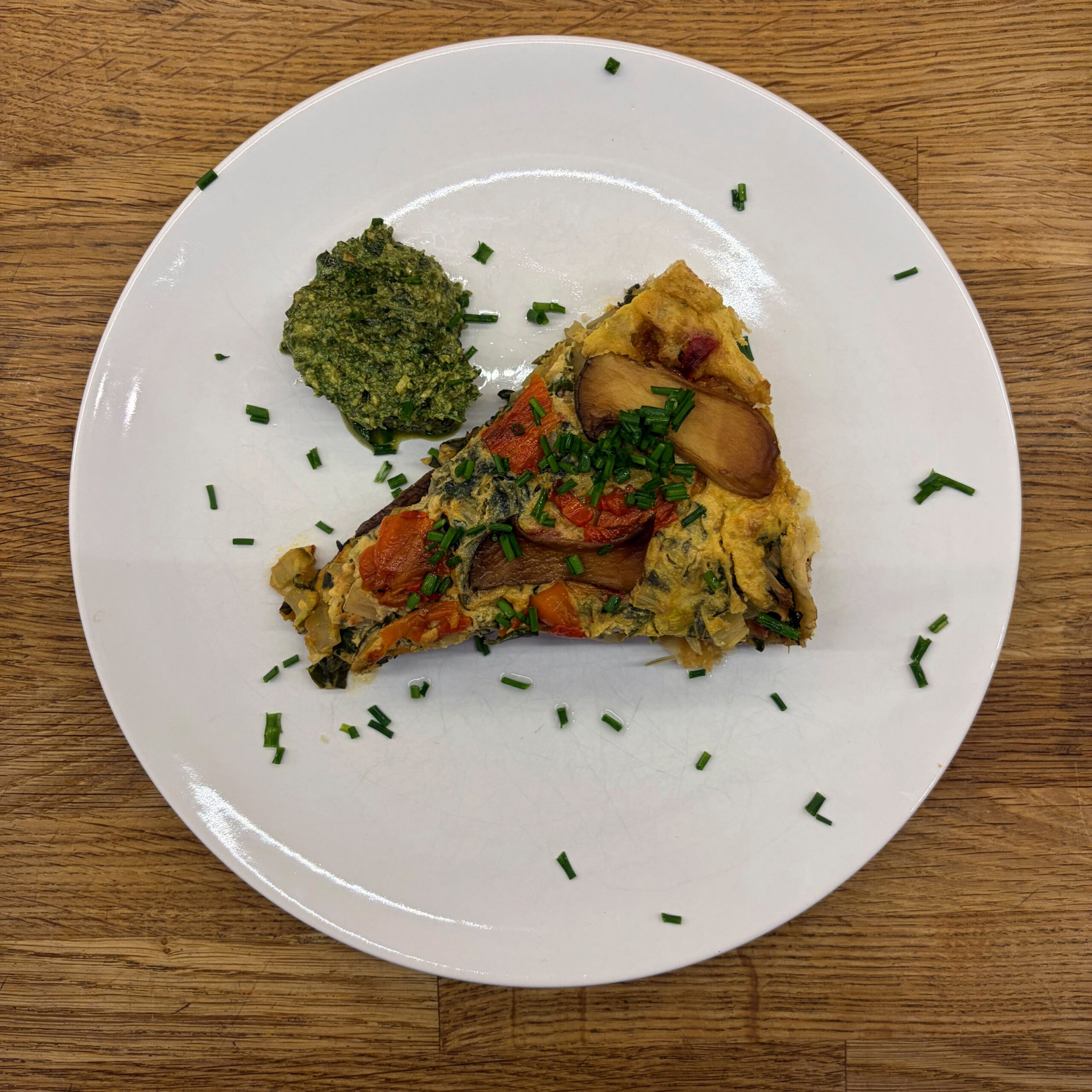 Ein Stück Mangold-Tarte mit Tomaten und Pilzen auf weißem Teller, garniert mit gehackten Schnittlauchröllchen, serviert mit einem Klecks Basilikum-Walnuss-Pesto.