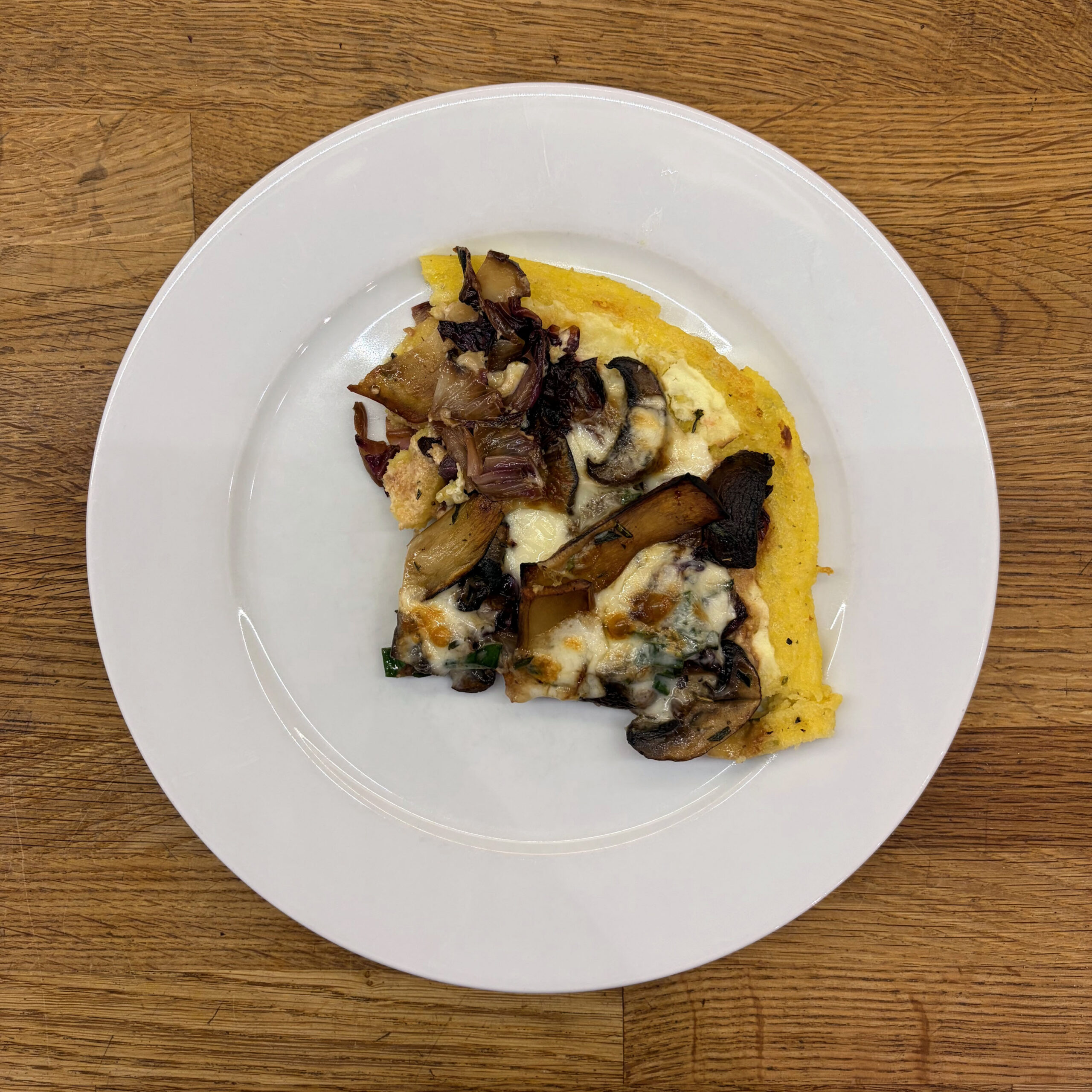 Ein Stück Polentapizza mit sautierten Champignons und Kräuterseitlingen, gebratenen Radicchiostreifen und geschmolzenem Taleggio auf einem weißen Teller, von oben fotografiert.