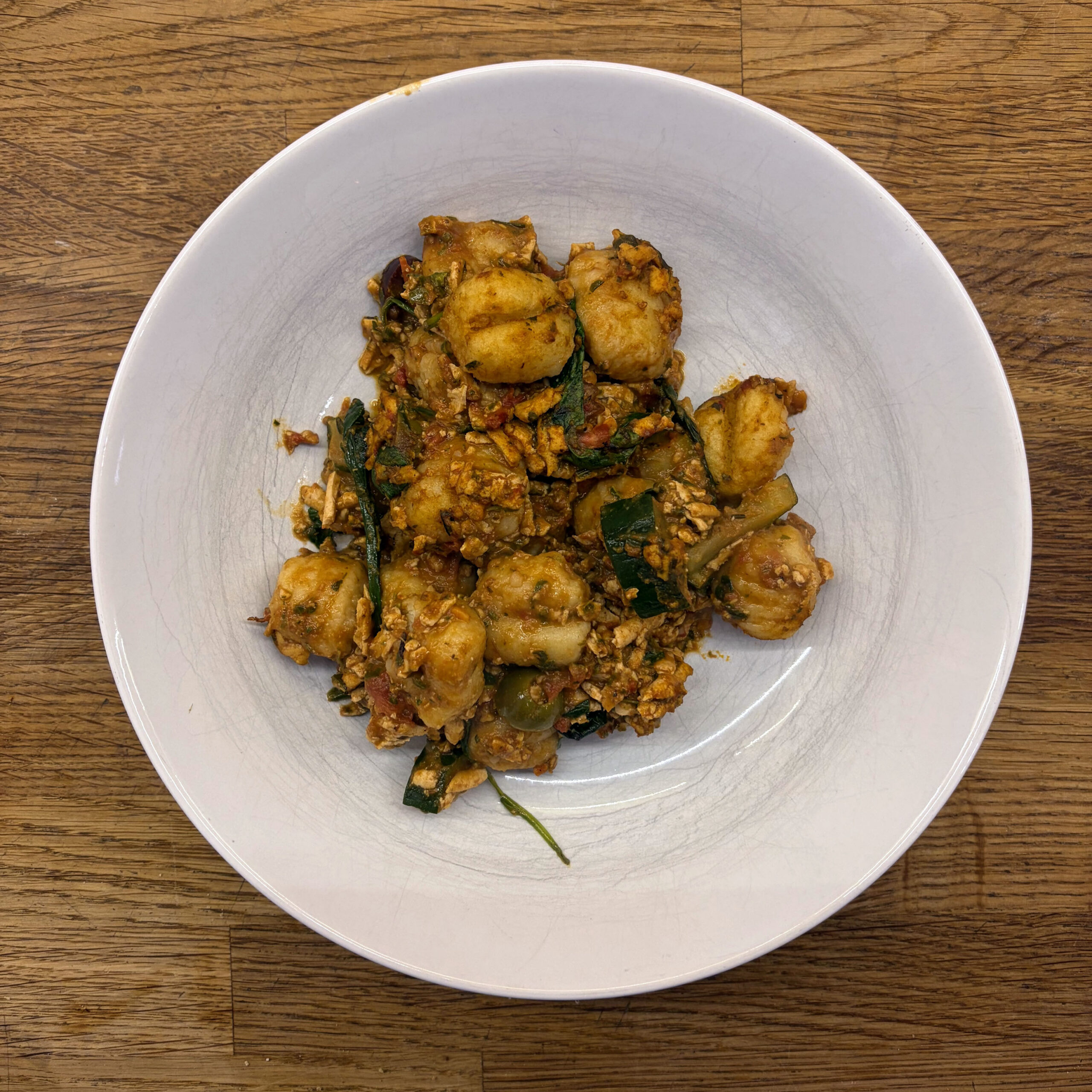 Weißer tiefer Teller mit gebackenen Gnocchi in würziger Tomatensoße, durchzogen von gebratenen Tofuwürfeln, Zucchinistücken und frischem Basilikum.