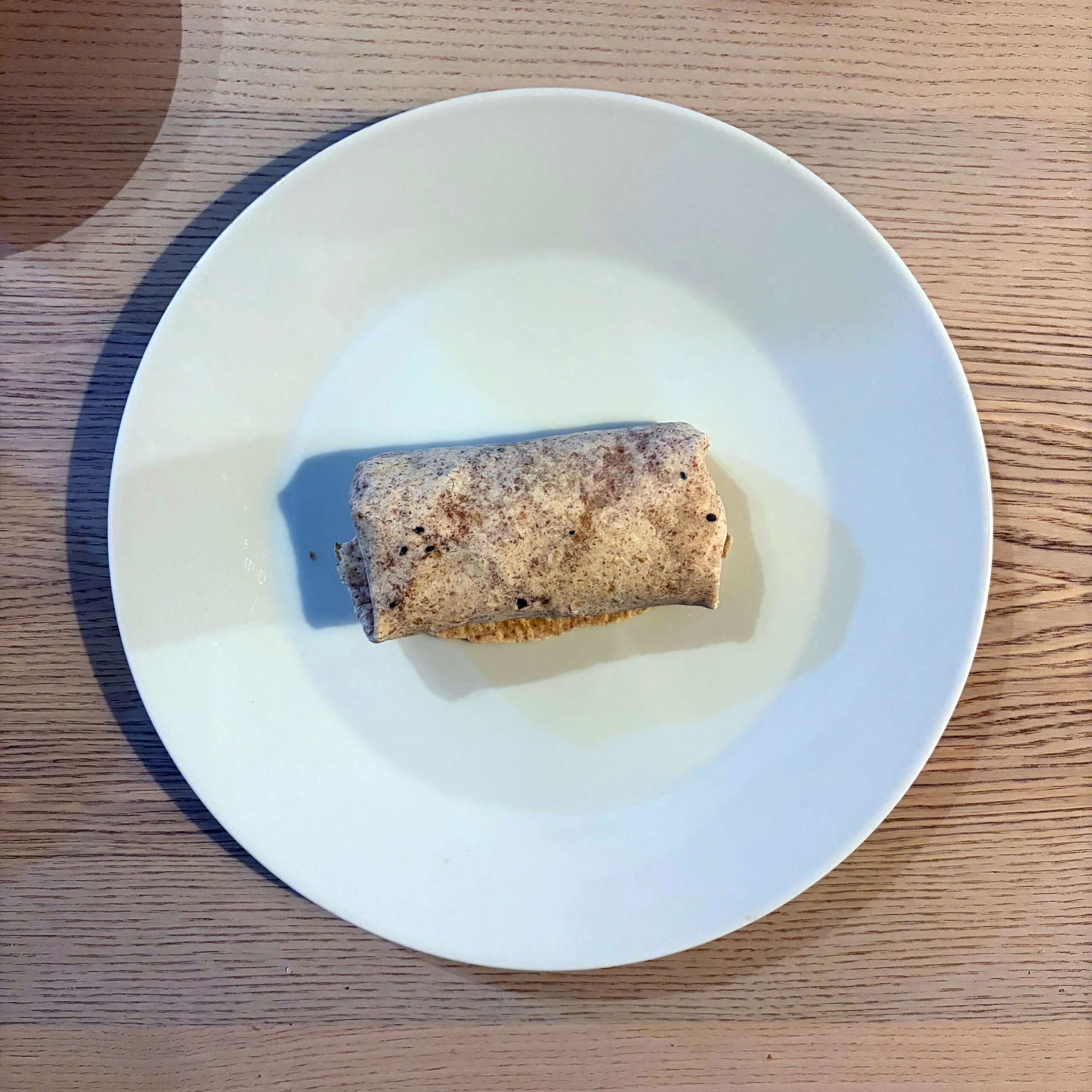 Ein einzelner, kompakt gerollter Wrap mit hellbraunem, dunkel gesprenkeltem Fladen liegt mittig auf einem großen weißen Teller; an einer Seite quillt etwas goldbraune Füllung hervor. Der Teller steht auf einer hellbraunen, längs gemaserten Holzfläche, aufgenommen aus leicht schräger Draufsicht.