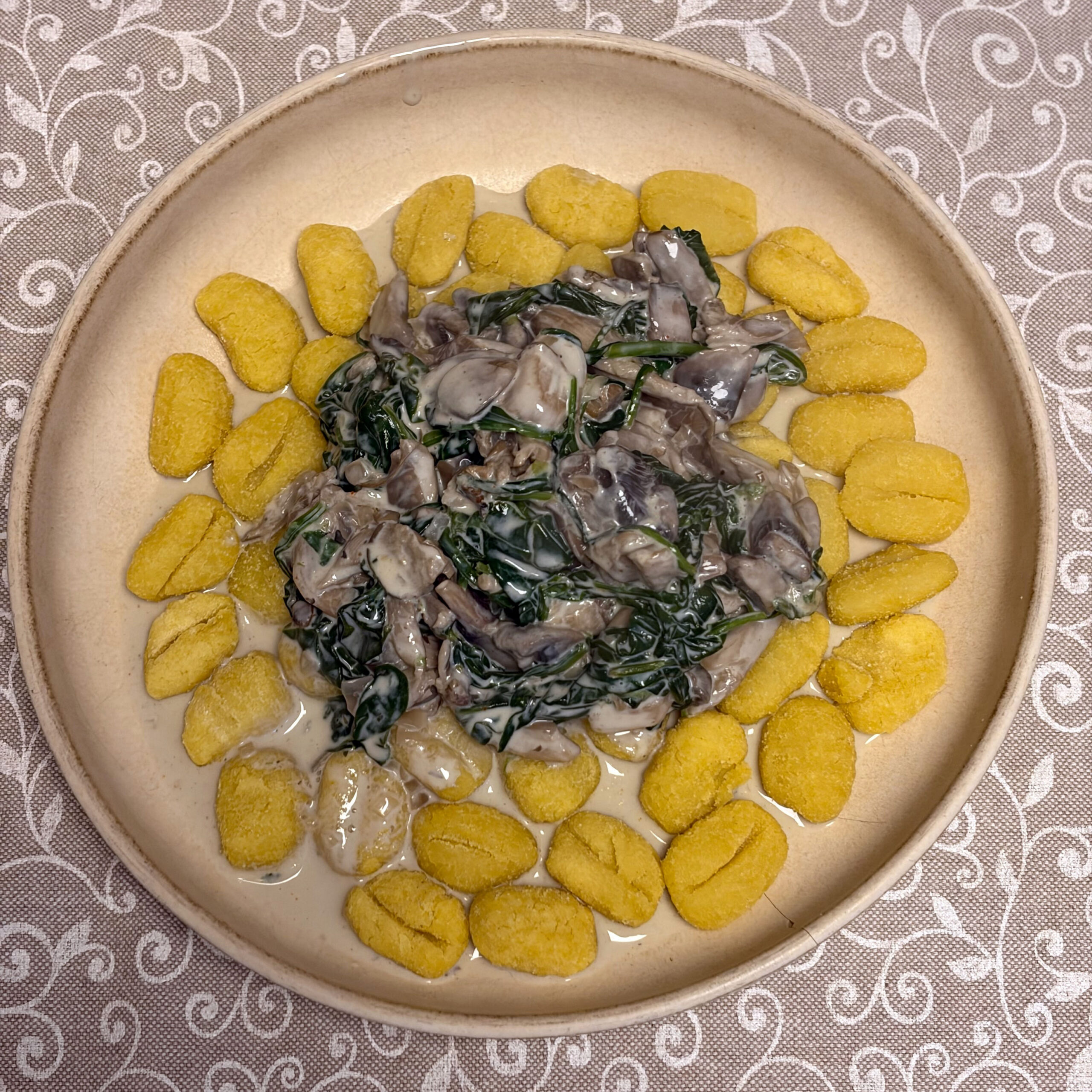 Goldgelbe, leicht gebräunte Gnocchi liegen ringförmig auf einem großen, beigefarbenen Keramikteller. In der Mitte befindet sich eine cremige, helle Soße mit graubraunen Pilzstücken und dunkelgrünem Spinat; der Teller steht auf einer grau-weiß gemusterten Stoffdecke, fotografiert von oben.